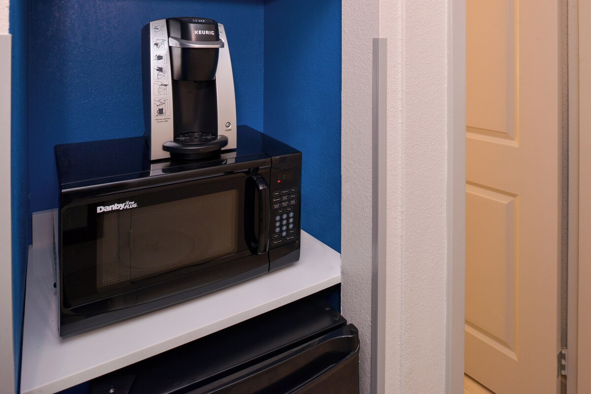 A microwave oven with a coffee maker on top, placed on a white shelf in a room with blue walls.