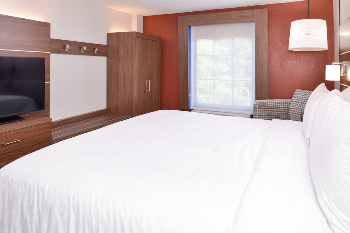 A hotel room with a large bed, a TV on a wooden cabinet, a window with blinds, and a lamp on the wall.
