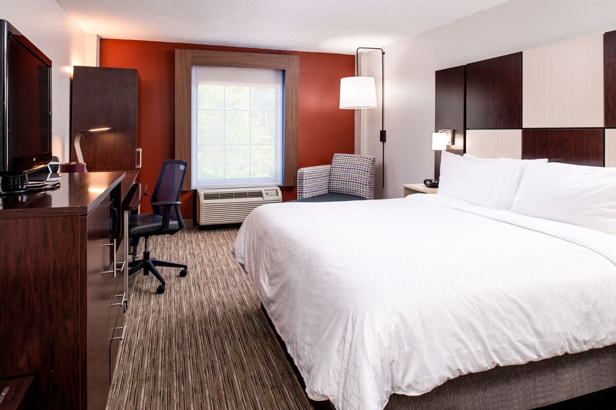 A hotel room with a large bed, a desk with a chair, a television, and a window with a view of trees.