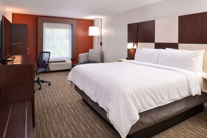 A hotel room with a large bed, a desk with a chair, a television, and a window with blinds.