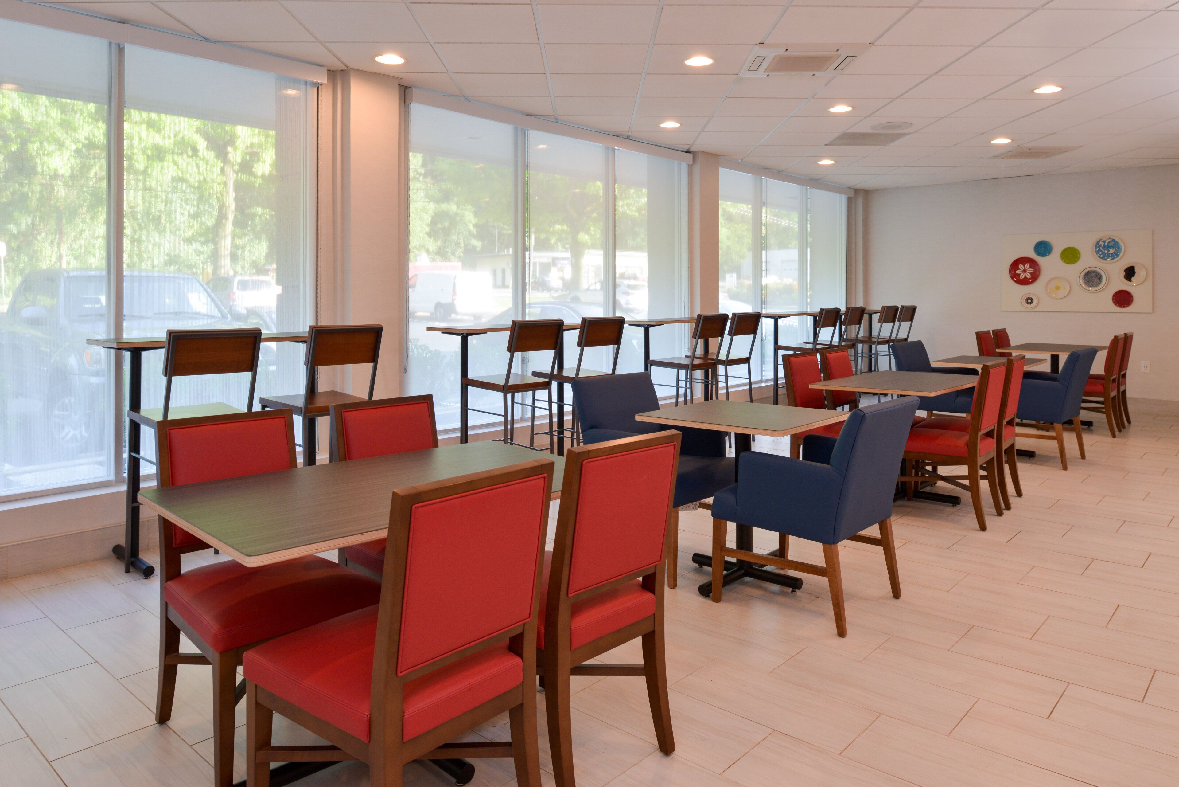 A spacious indoor dining area with multiple tables and chairs, large windows offering a view of the outside, and a wall decorated with circular plates.