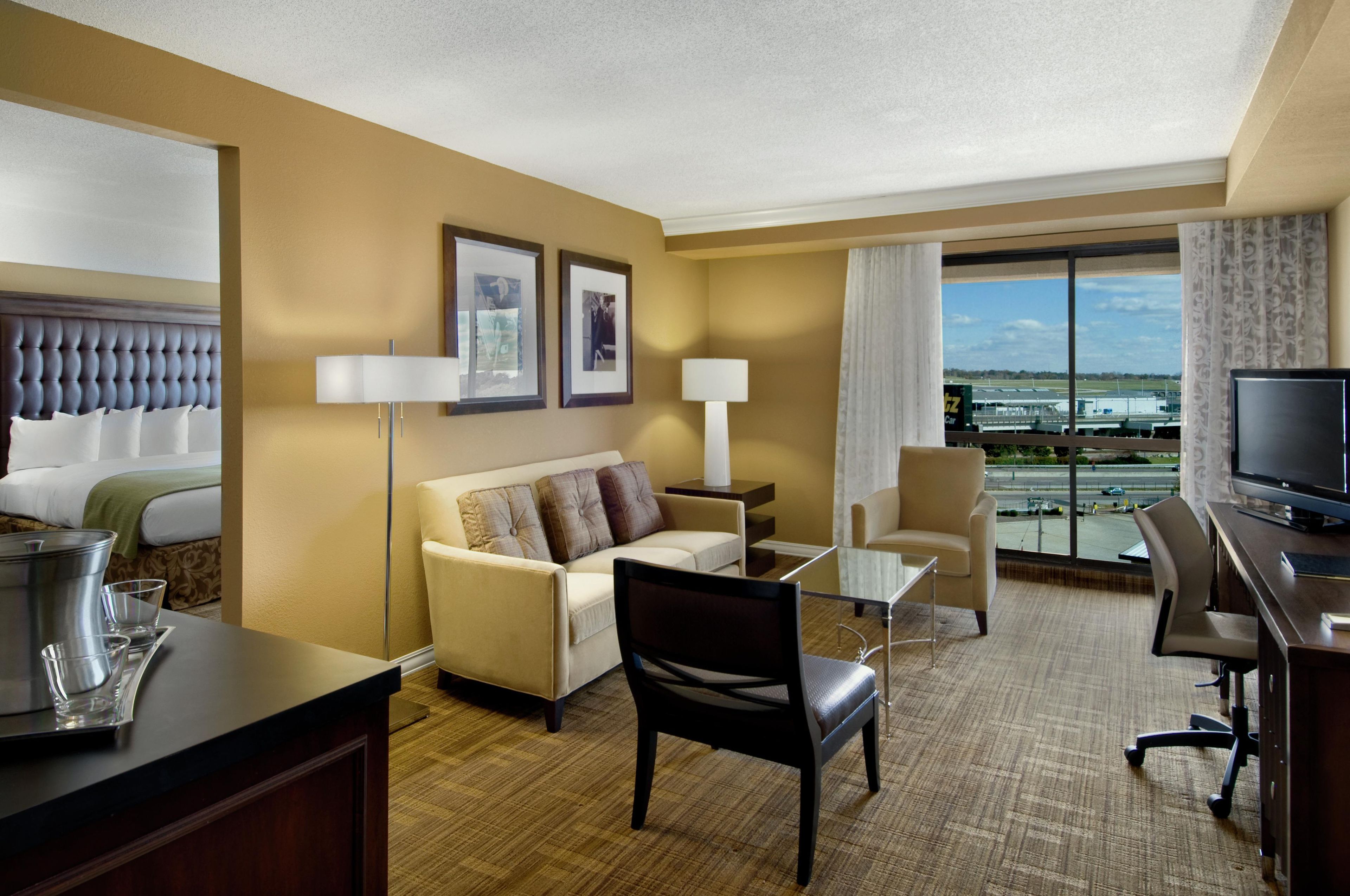 A hotel room with a bed, a sofa, a desk, a TV, and a view of the airport.