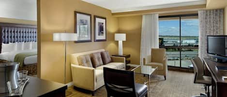A hotel room with a bed, a sofa, a desk, a TV, and a view of the airport.
