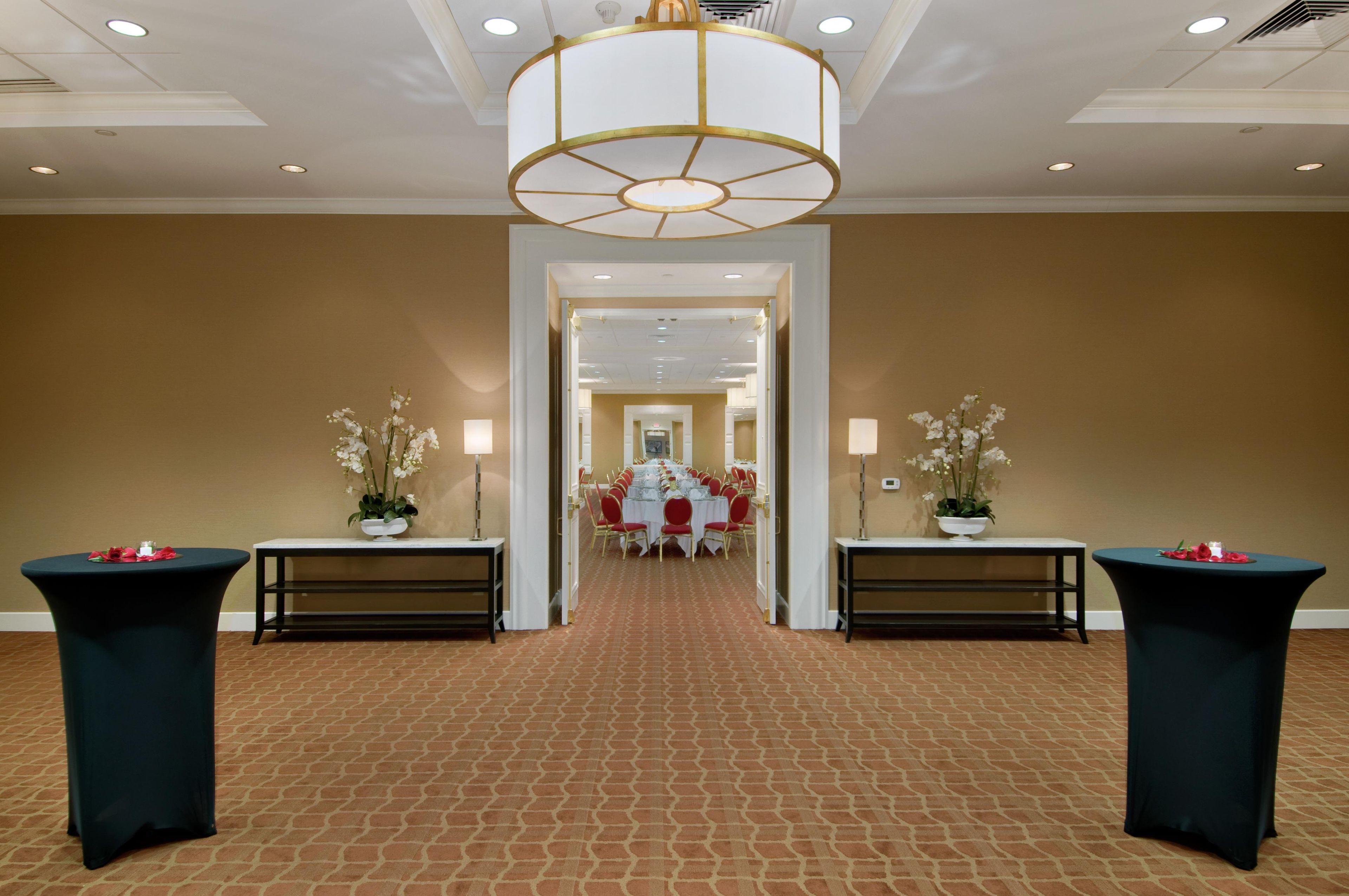 A hotel corridor with a carpeted floor, a chandelier, tables with potted plants, and a dining area in the distance.