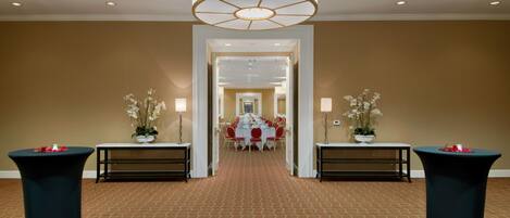 A hotel corridor with a carpeted floor, a chandelier, tables with potted plants, and a dining area in the distance.