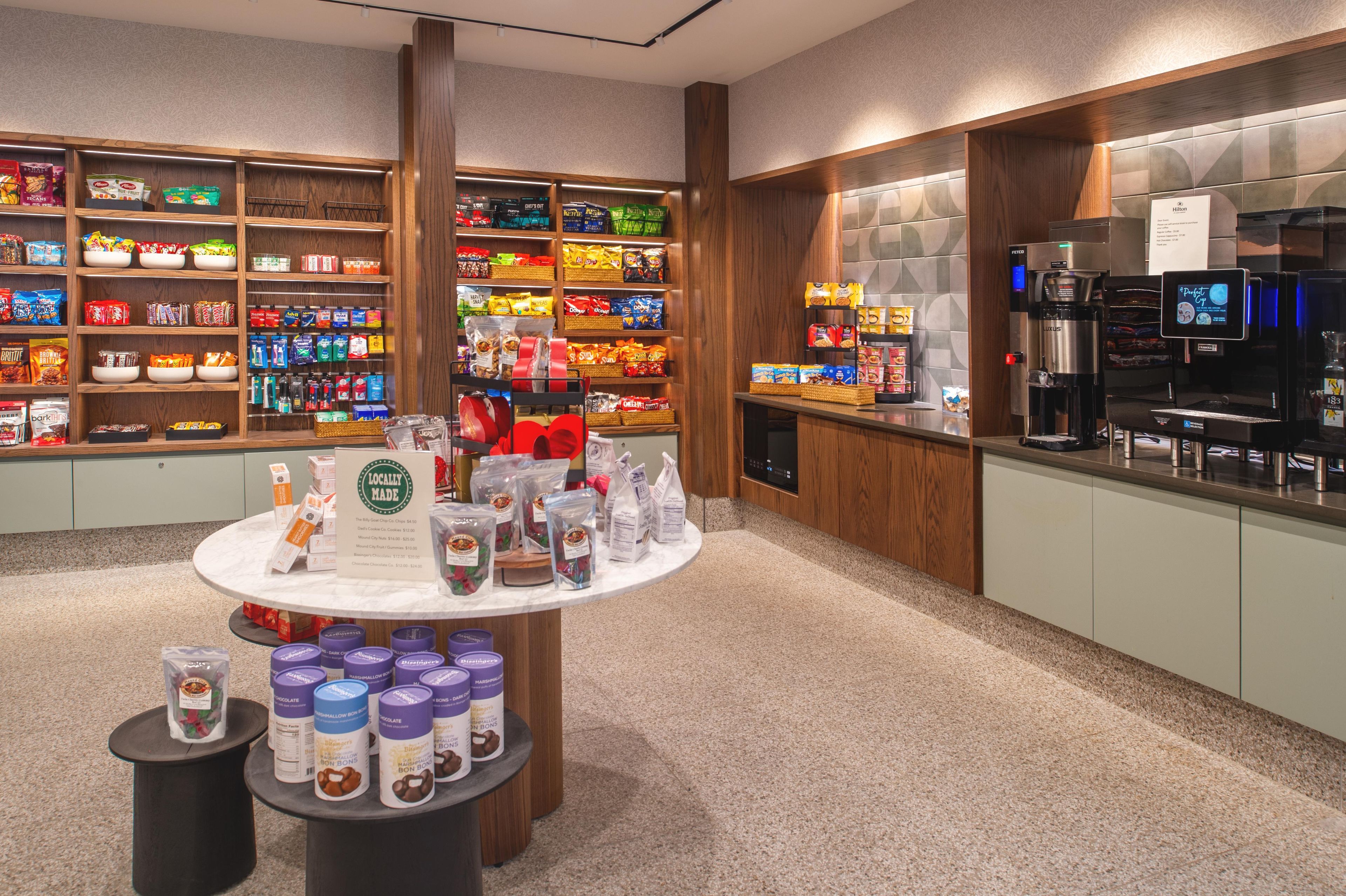 A hotel lobby with a self-service beverage station, a display of locally made snacks, and a variety of packaged goods on shelves.
