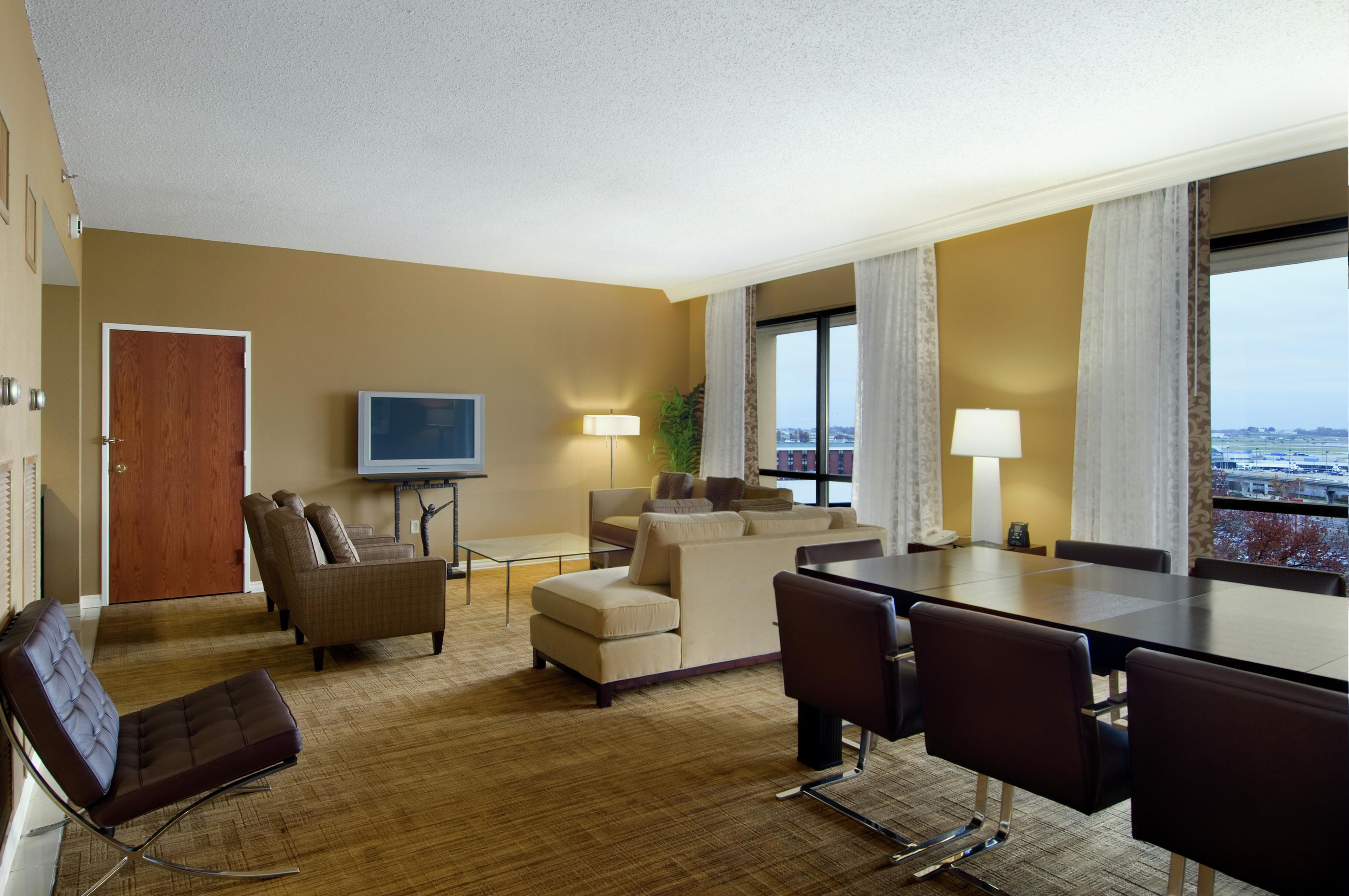 A hotel room with a sofa, chairs, a dining table, a TV, and a large window with a view.