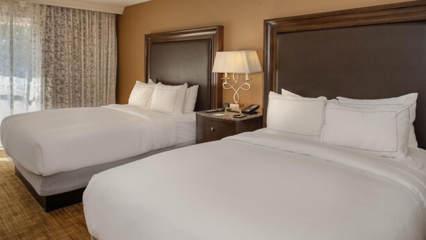 A hotel room with two beds, a nightstand with a lamp, and a window with curtains. - Hilton St. Louis Airport (St. Louis)