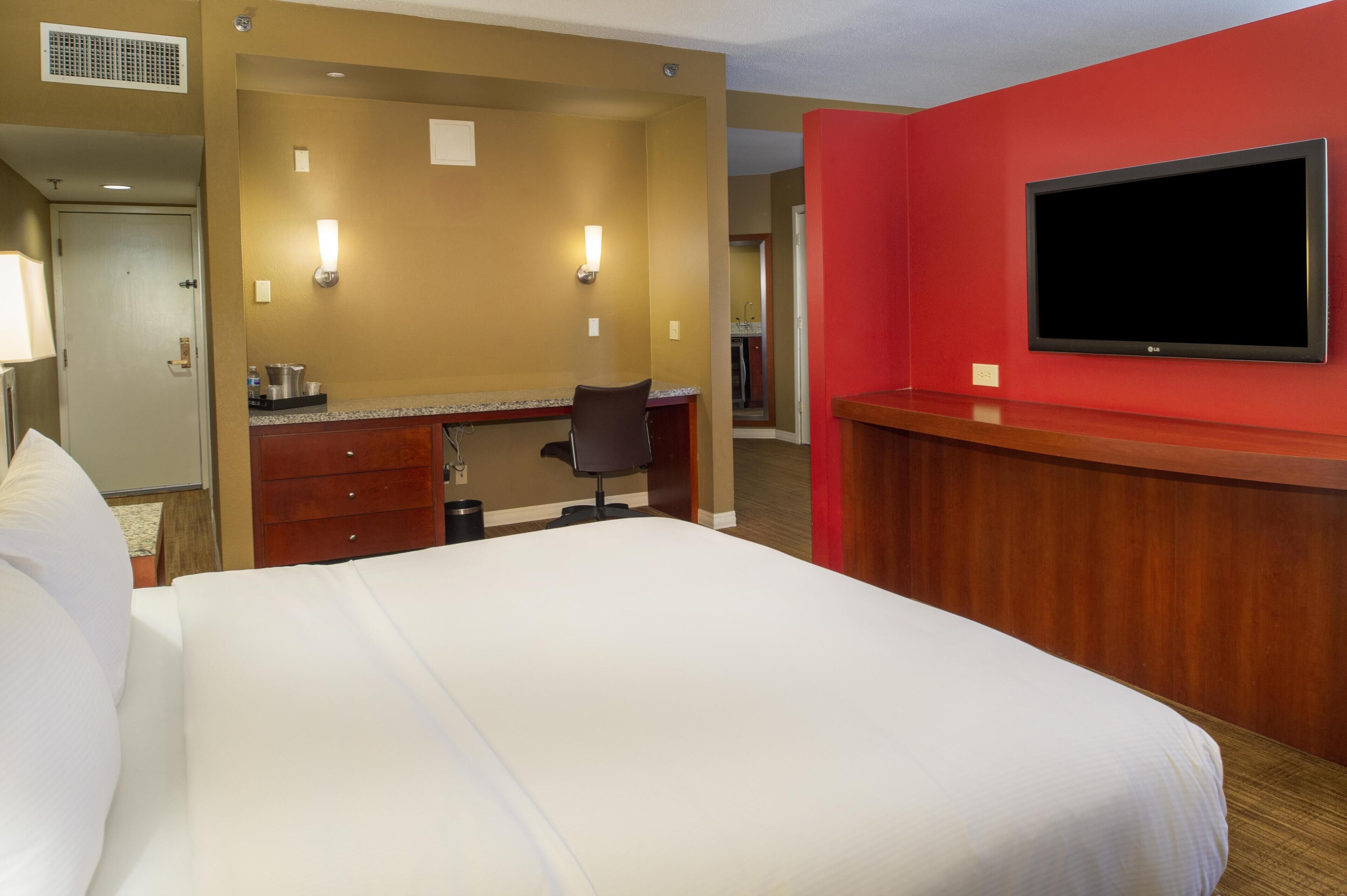 A hotel room with a bed, a desk with a chair, a television, and a bathroom visible in the background.