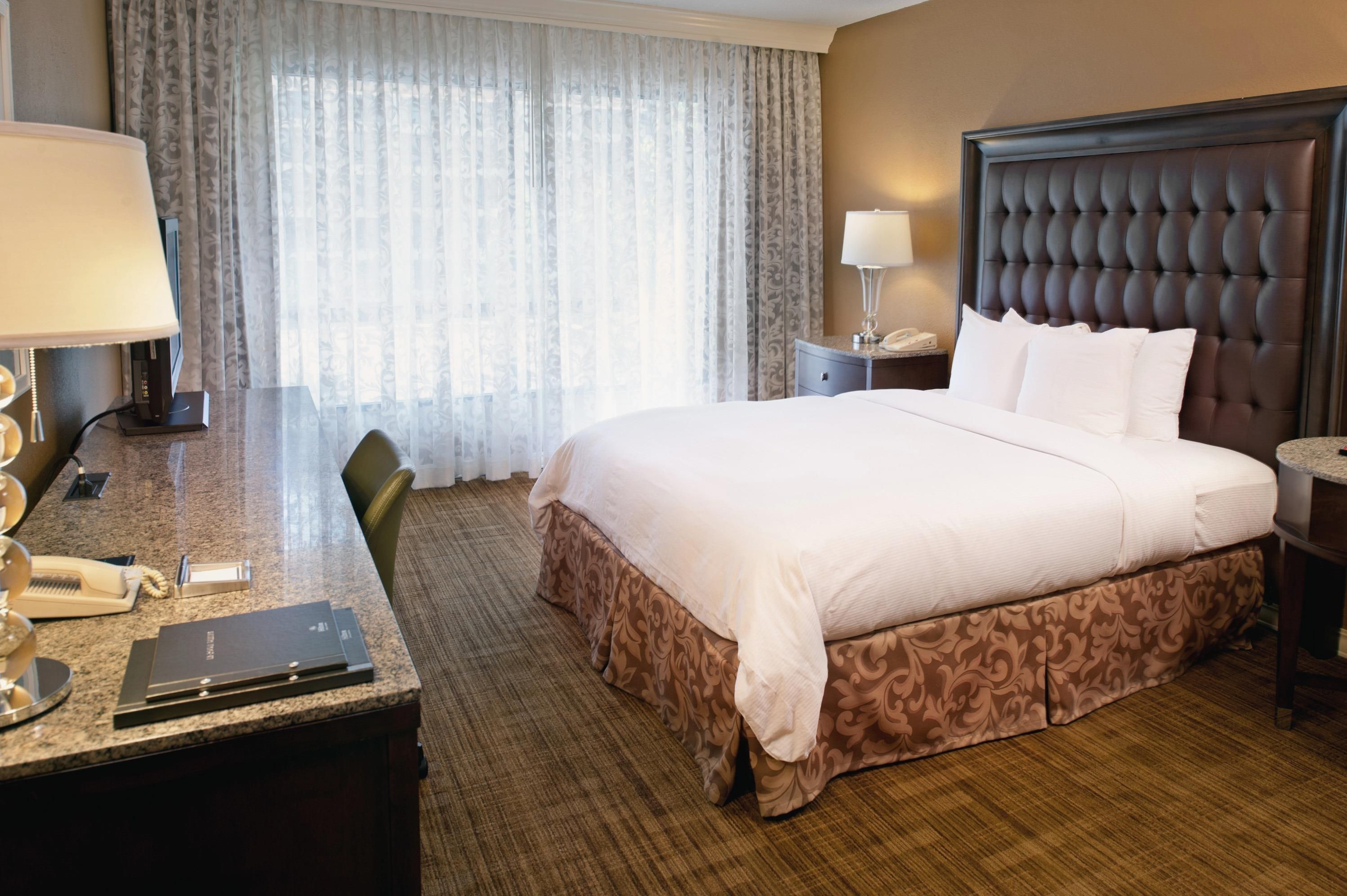 A hotel room with a large bed, a desk with a lamp, a telephone, and a window with curtains.