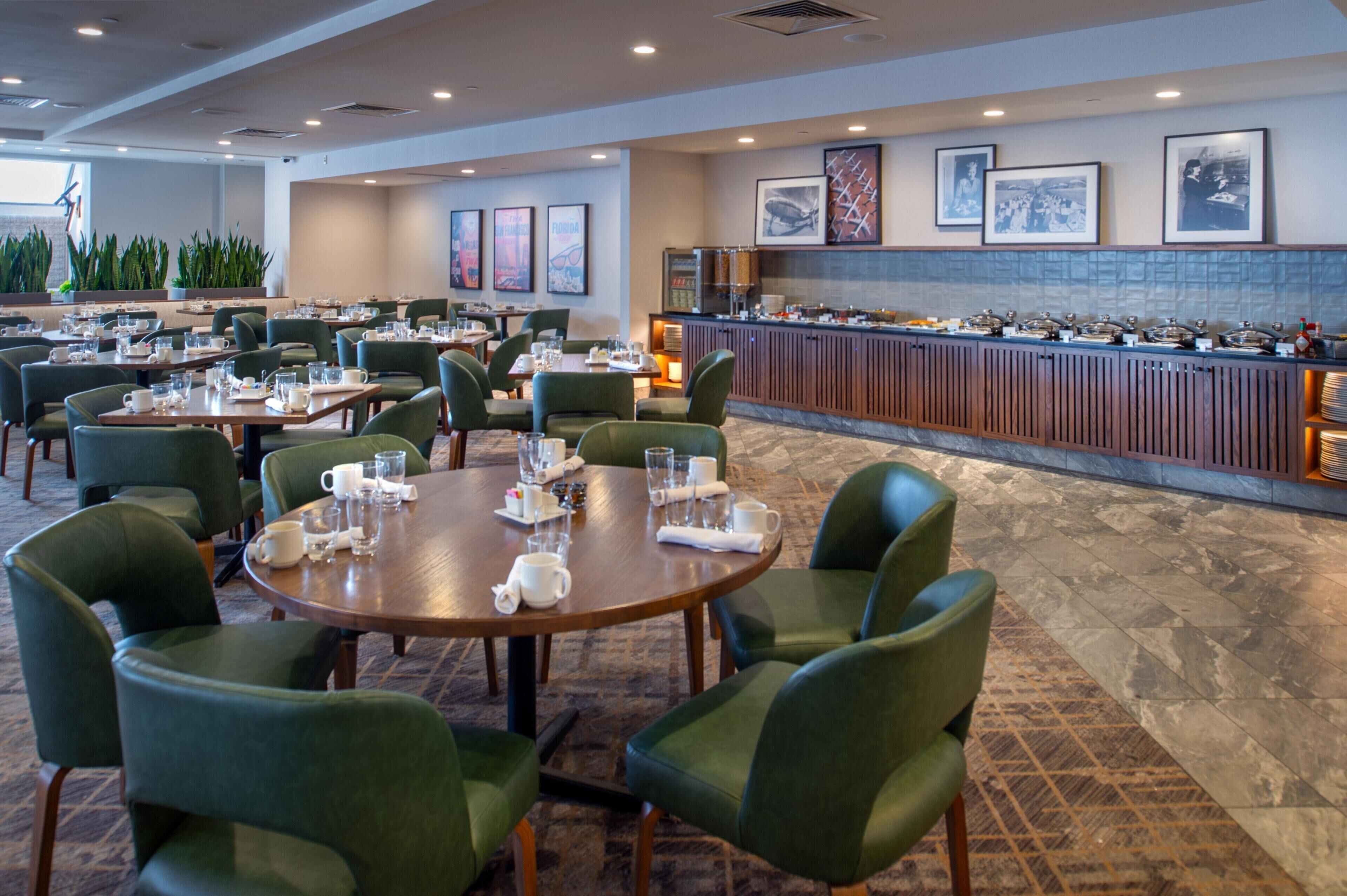 A hotel dining area with round tables, green upholstered chairs, a buffet bar, and framed pictures on the wall.