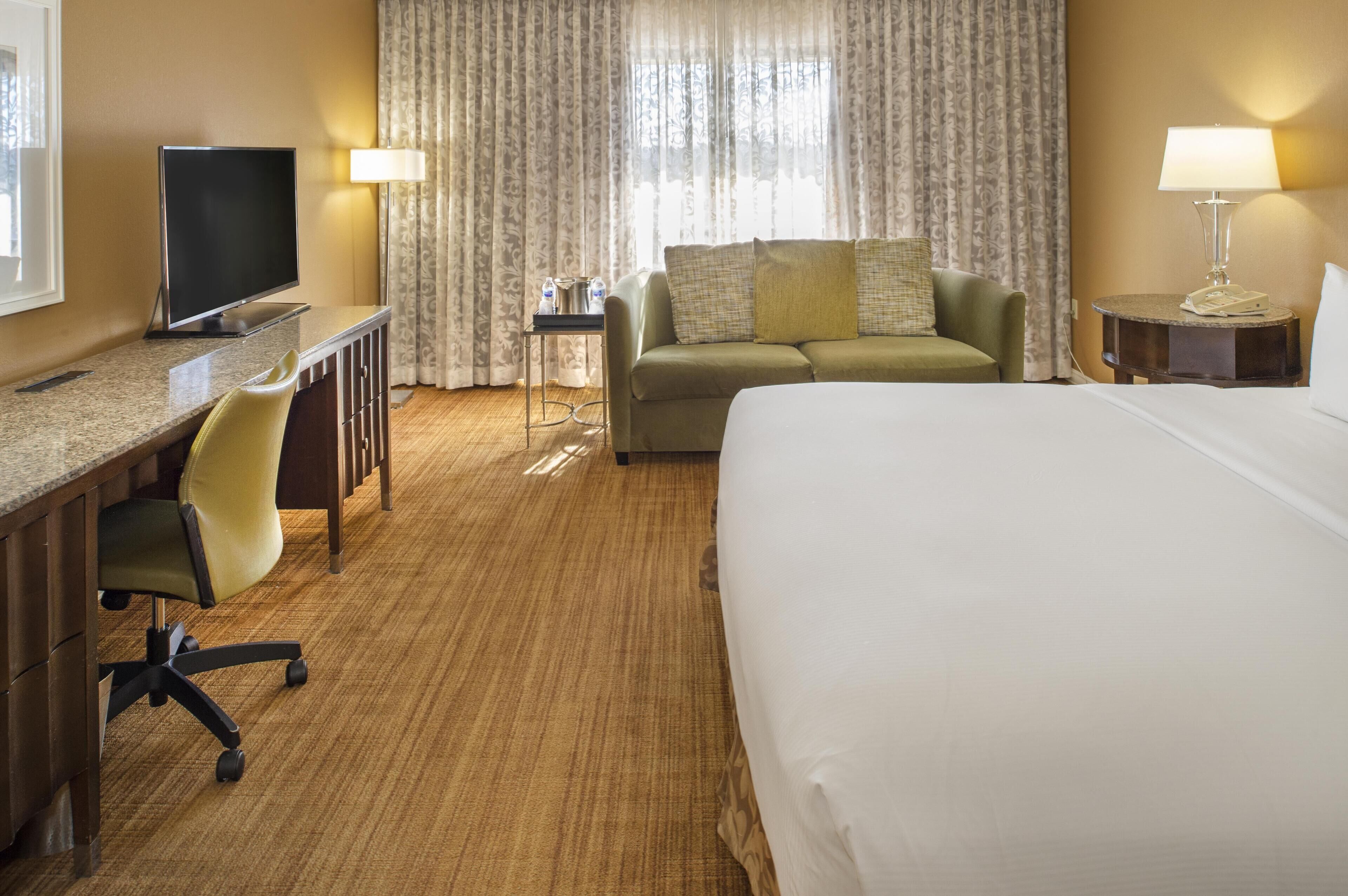 A hotel room with a bed, a desk with a chair, a television, a sofa, and two lamps.