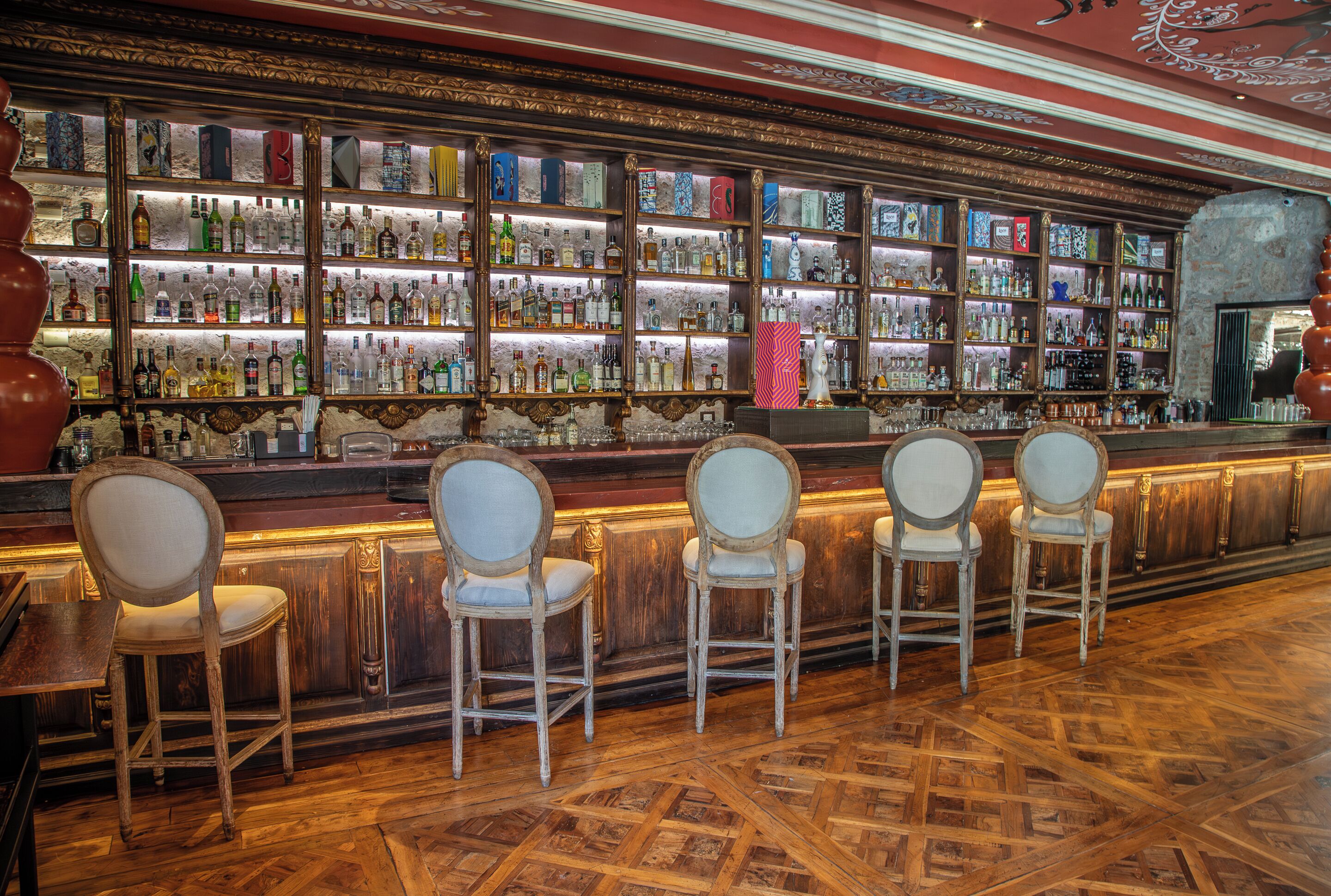 Um bar com várias prateleiras de garrafas, cadeiras de madeira e piso estampado.