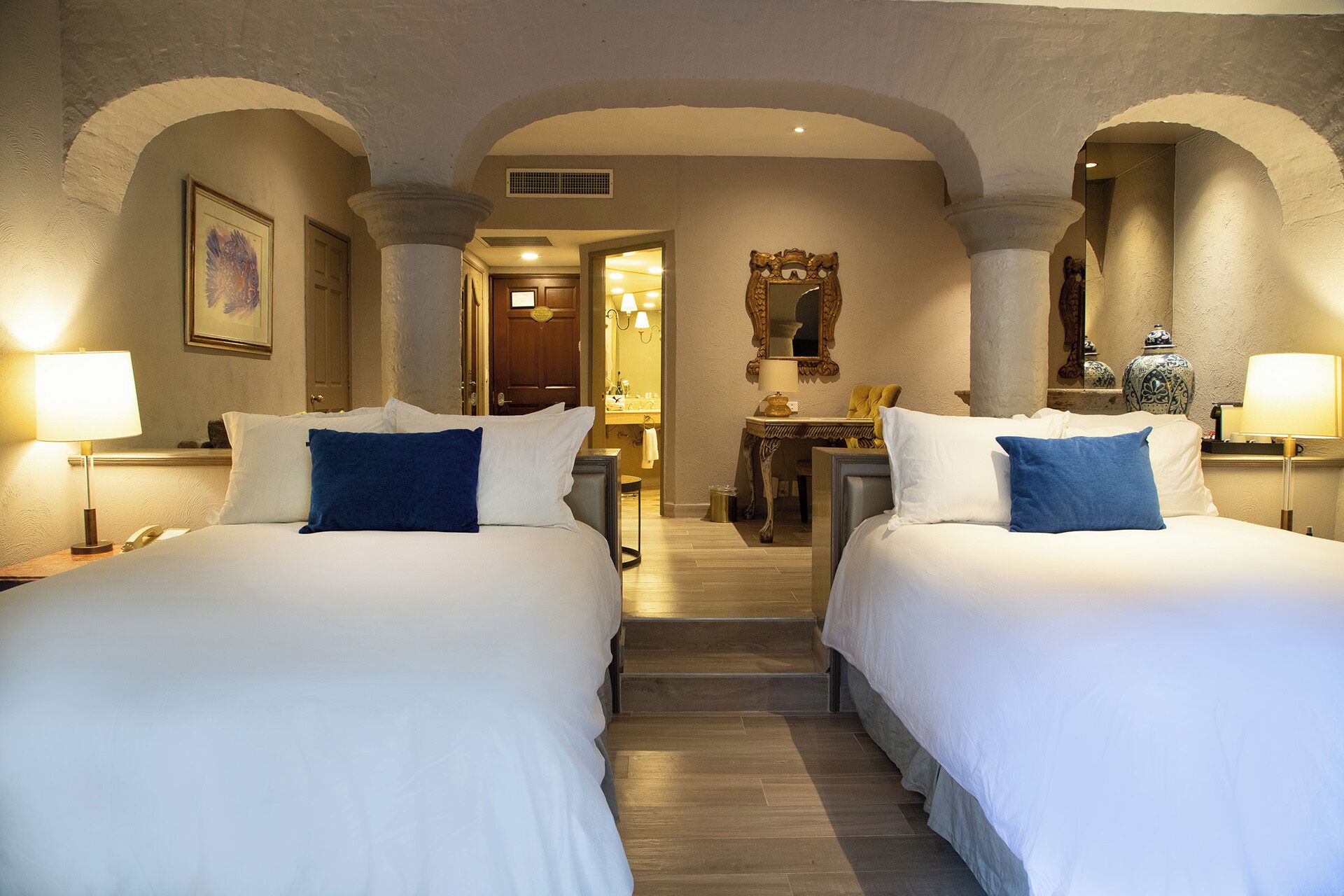 A hotel room with two beds, a desk, a mirror, and a bathroom visible through an archway.