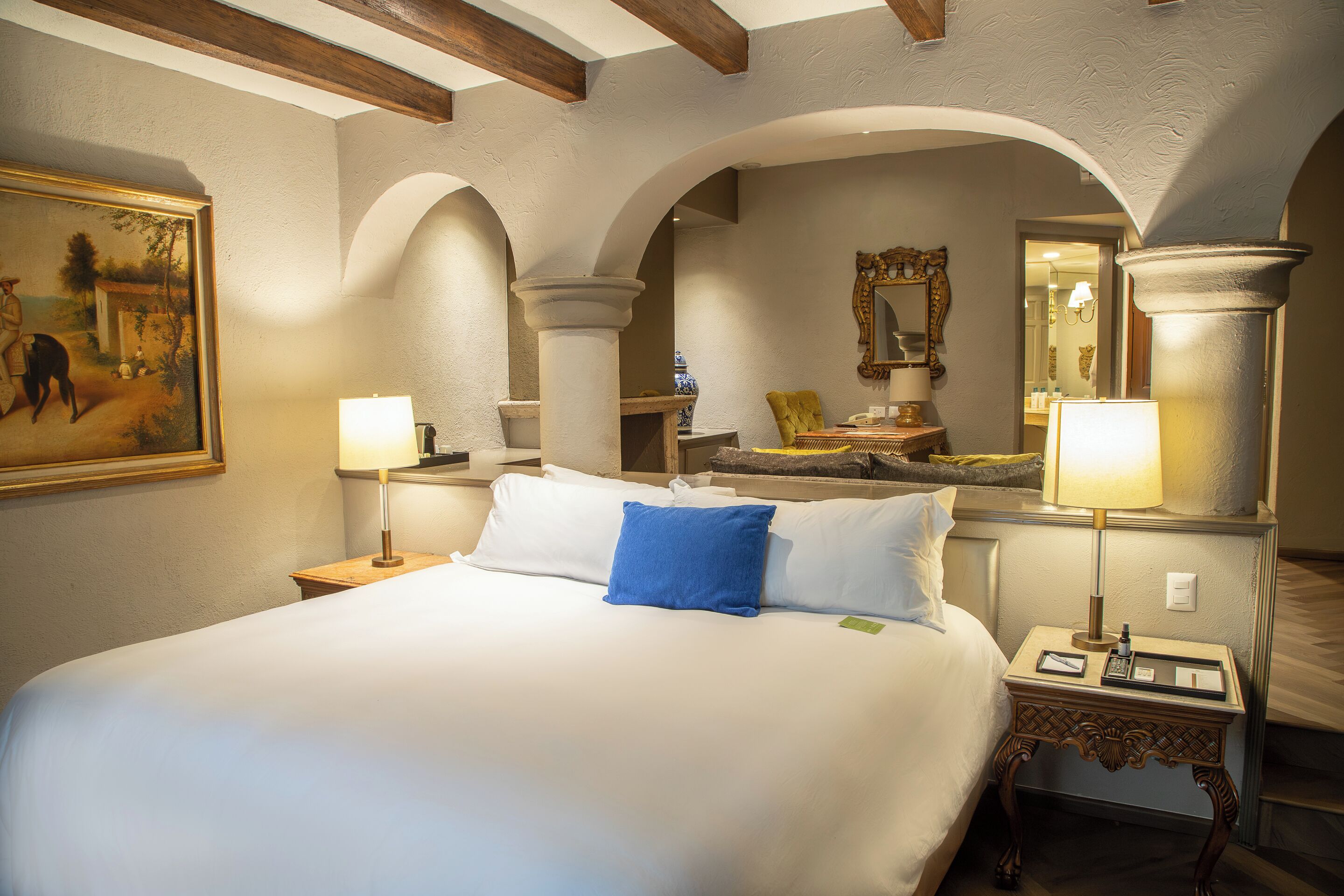 A hotel room with a large bed, two bedside tables, a nightstand, a mirror, and a painting on the wall.