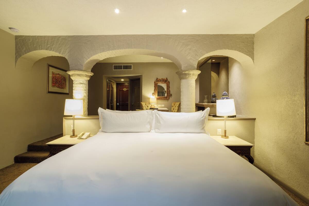 A large bed with two pillows, flanked by nightstands with lamps and a phone. There are three arches in the background, and a mirror above the bed.
