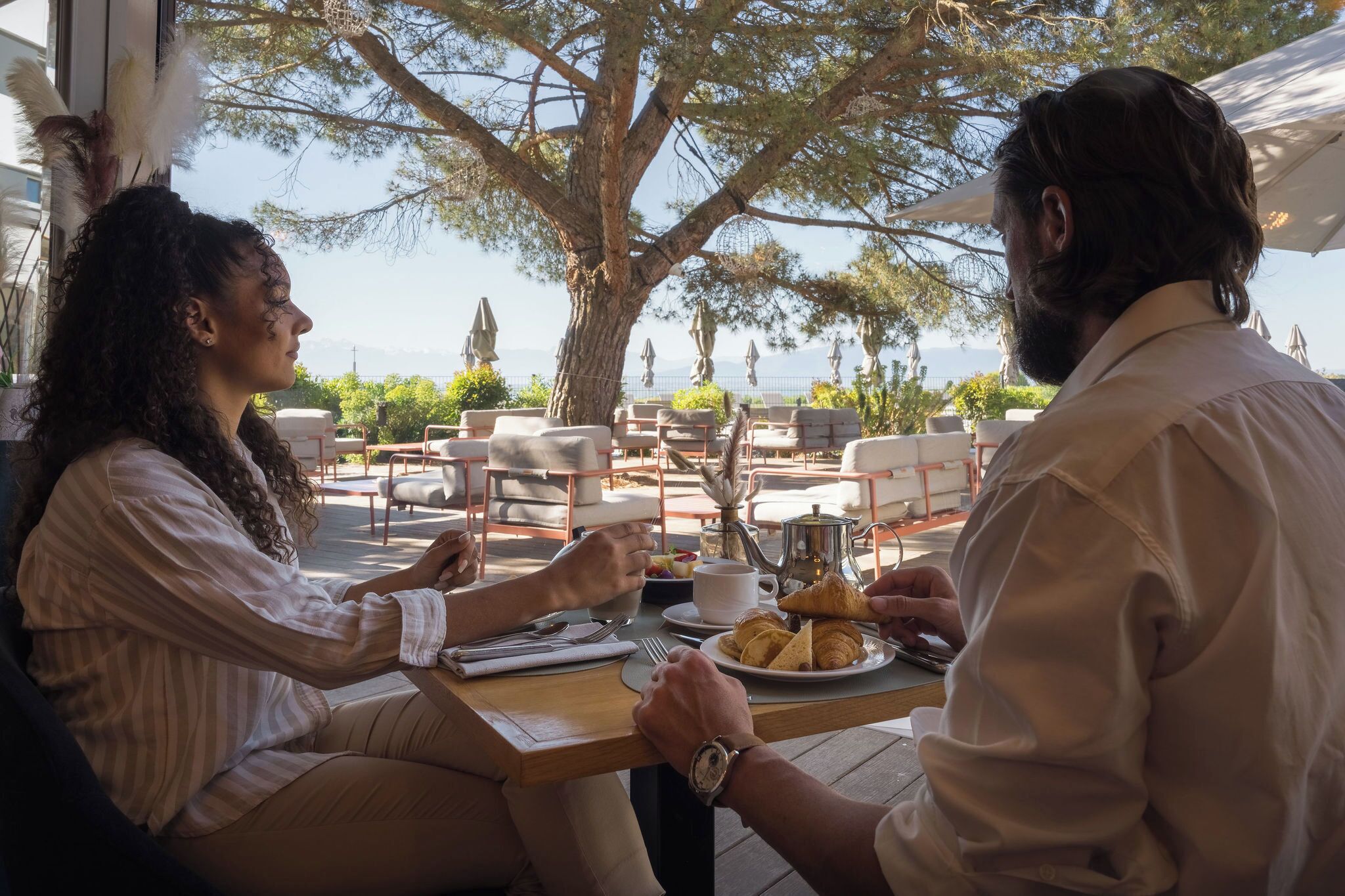 Ein Paar speist in einem Straßencafé mit Blick auf das Meer, Tische, Stühle und Sonnenschirme sind vorhanden.