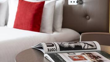 Una cama bien hecha con un cabecero gris, almohadas blancas y rojas, y una revista sobre la mesita de noche.