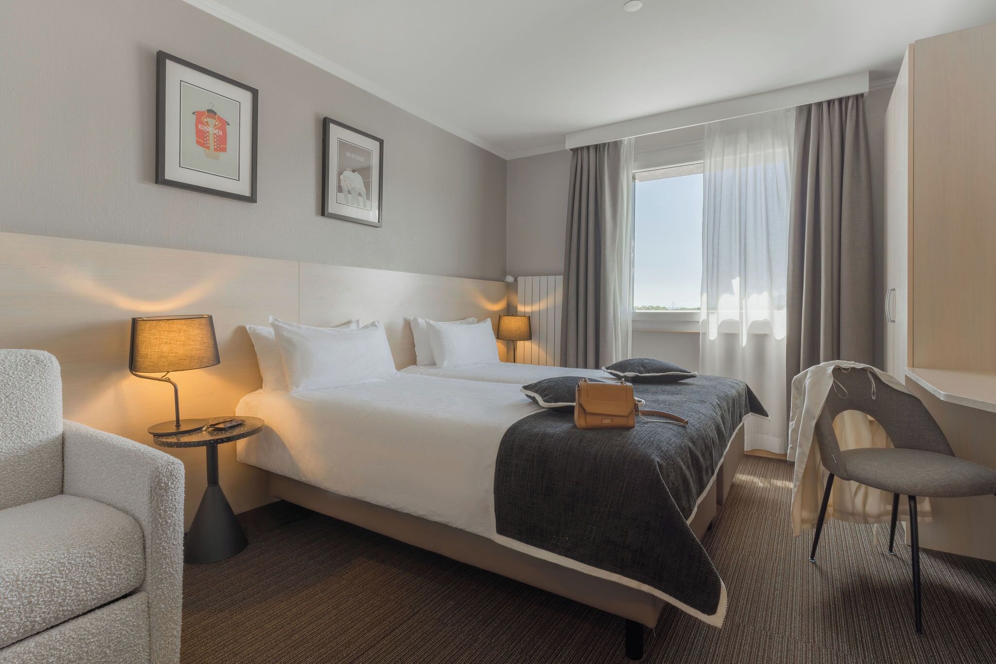 Une chambre d’hôtel avec un lit, deux lampes de chevet, une chaise, un bureau et une fenêtre avec des rideaux.