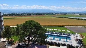 Un espace piscine agrémenté de chaises longues et de parasols, entouré d’un jardin paysager et offrant une vue sur un vaste champ et des montagnes au loin.