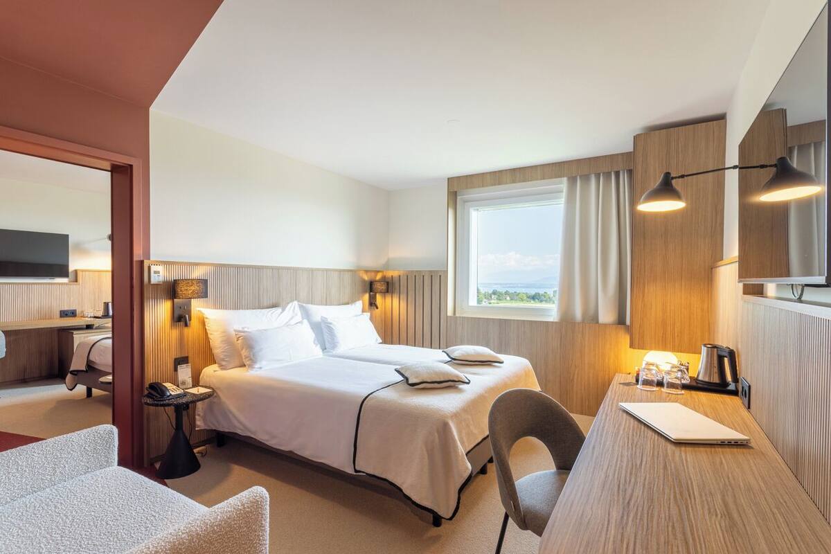 Habitación de hotel con cama, escritorio con computadora portátil, silla, mesita de noche y ventana con cortinas.