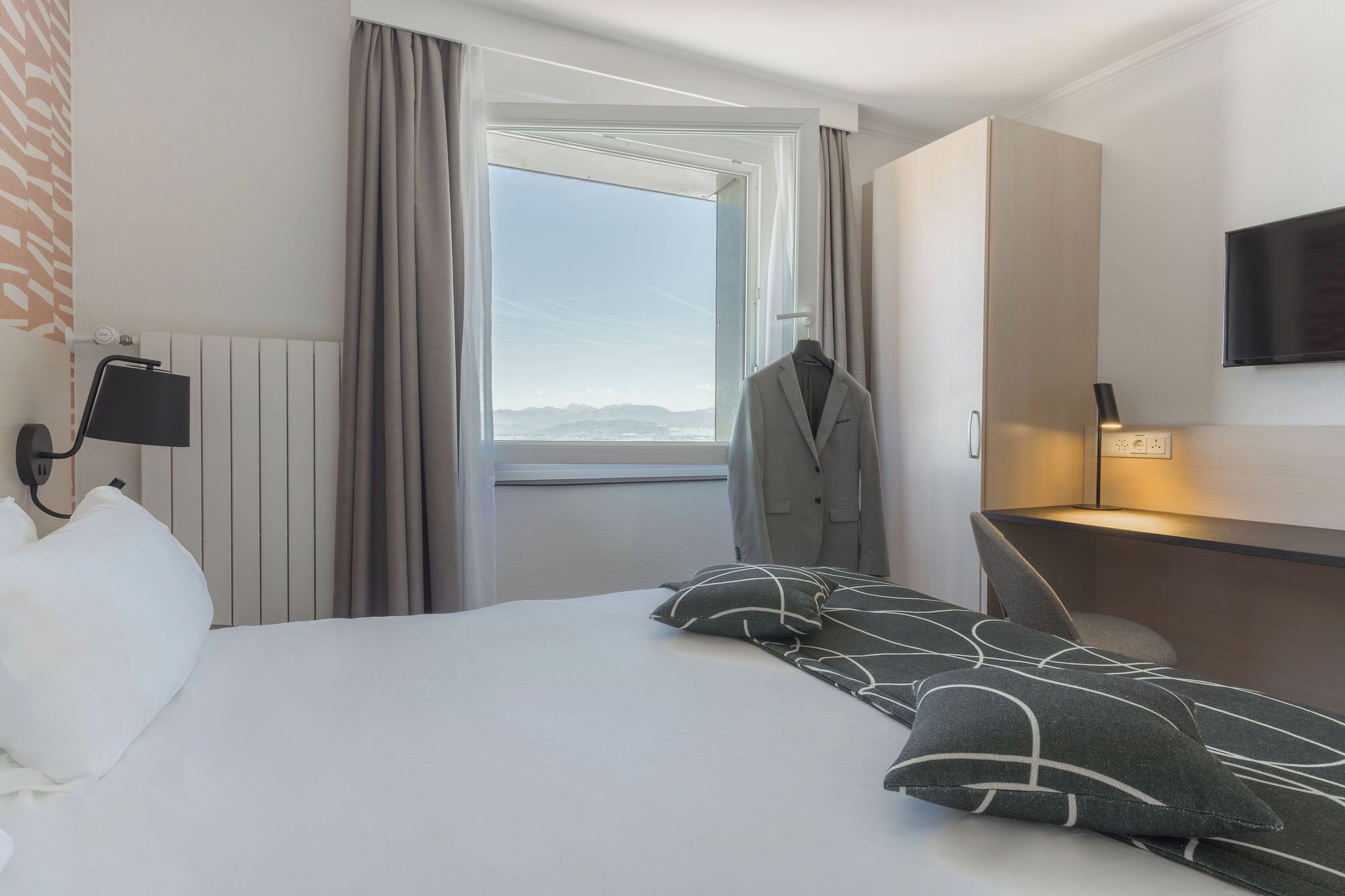 Ein Hotelzimmer mit Bett, Schreibtisch, Stuhl, Kleiderhaken, Fernseher und einem Fenster mit Vorhängen.