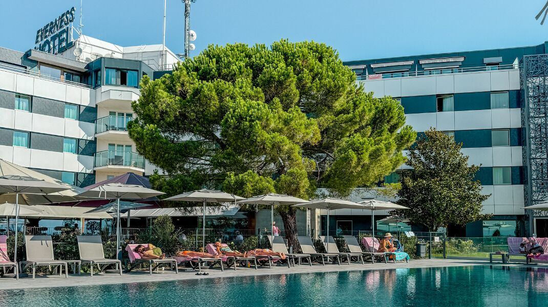 Un área de piscina hotelera con sillones de descanso y sombrillas, un árbol grande y un edificio con el letrero "Everness Hotel".