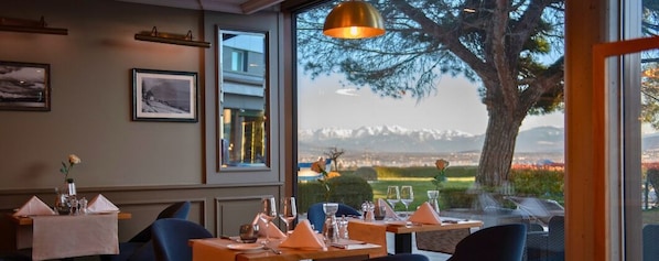 Un restaurante con mesas dispuestas para comer, cuadros en la pared y vistas a un paisaje montañoso a través de amplias ventanas.