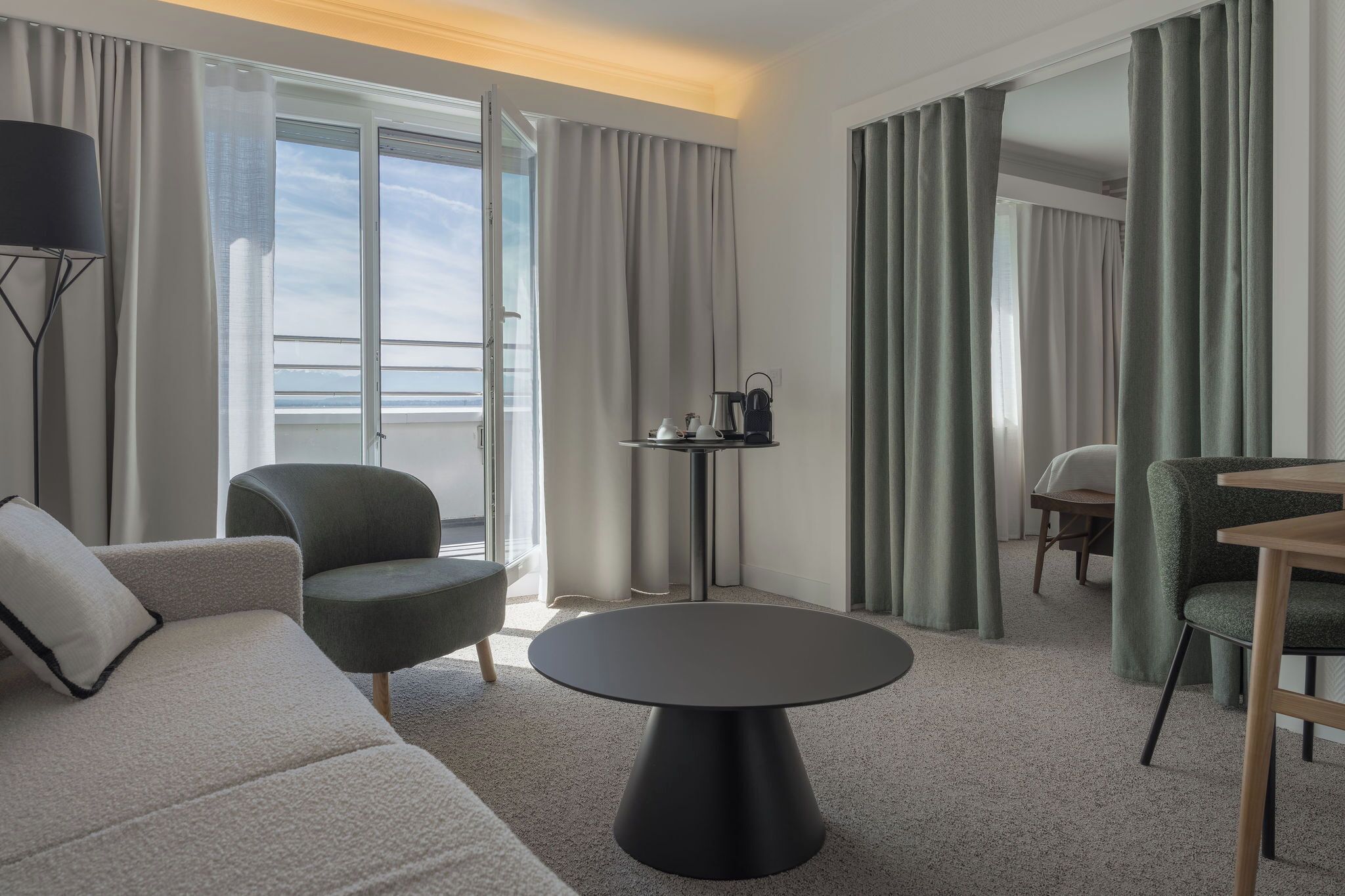 A hotel room with a sofa, armchair, round table, and a view of the sea through large windows.