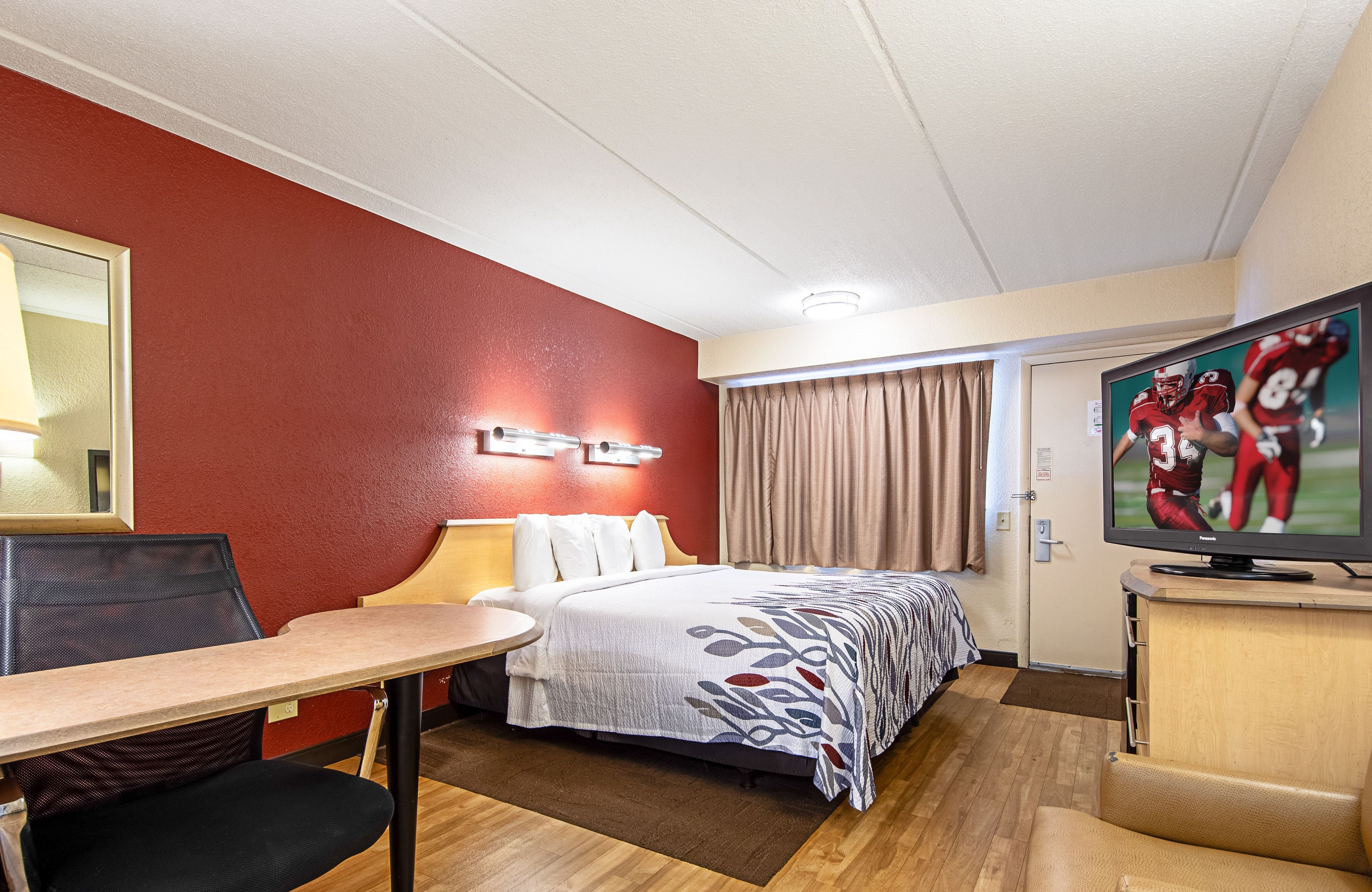 Habitación de hotel con cama, escritorio, silla, televisor y ventana con cortinas.