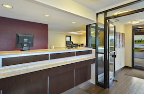A reception desk with a computer monitor, a cash register, and a telephone. There is a glass door leading to a hallway. - Red Roof Inn Charleston West - Hurricane, WV (Hurricane)