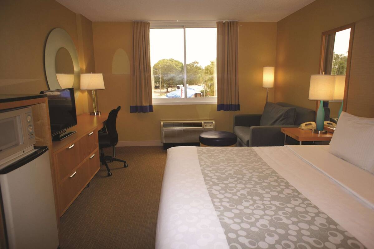 Ein Hotelzimmer mit Bett, Schreibtisch, Stuhl, Fernseher, Lampe und einem fenster mit Vorhängen.