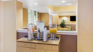 A breakfast bar with juice dispensers and a coffee machine, a plant, and a TV in the background. - La Quinta Inn & Suites by Wyndham Sarasota Downtown (Sarasota)