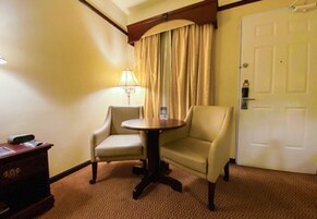 A hotel room with a round table, two armchairs, a lamp, a door, and a small desk with a telephone.