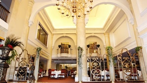 Un lobby imponente con techo alto, columnas ornamentales y una araña. Varias áreas de comedor con mesas dispuestas para los huéspedes.