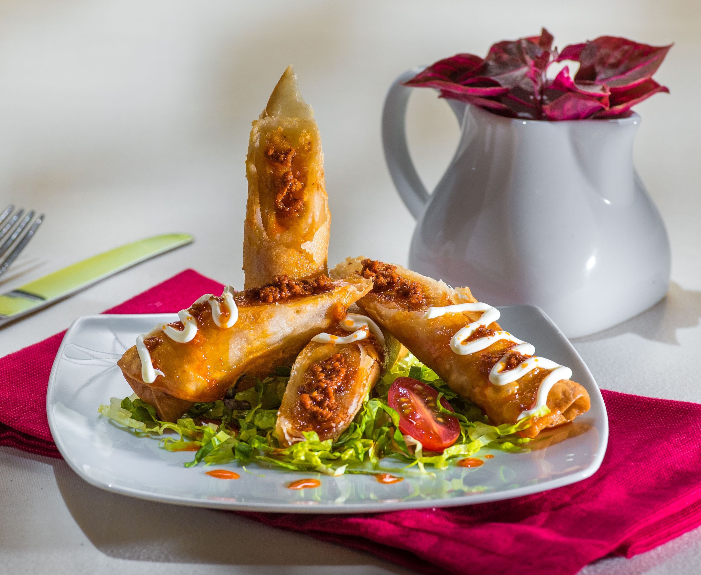 A plate of spring rolls with lettuce and a dipping sauce, accompanied by a pitcher of water and a fork on a table.