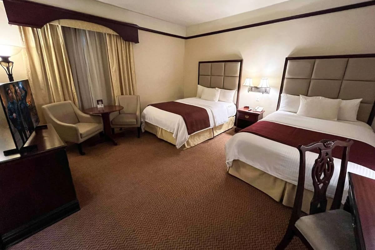 A hotel room with two beds, a desk, a chair, and a lamp.