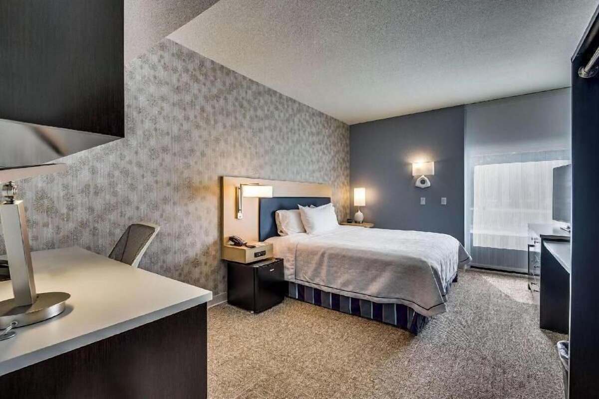 A hotel room with a bed, bedside table, lamp, desk with computer, and a chair.