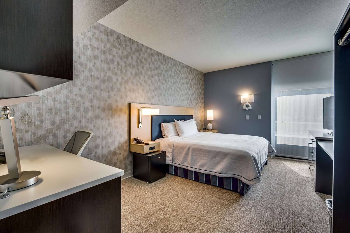 A hotel room with a bed, bedside table, lamp, and a desk with a chair.