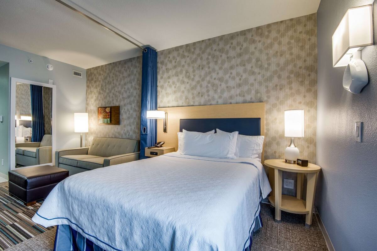 Una habitación de hotel con una cama grande, dos mesitas de noche, una silla, un escritorio y un espejo.