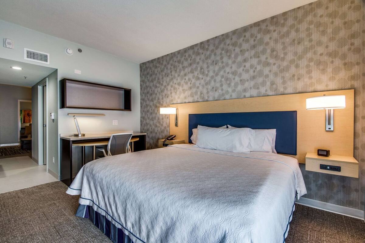 A hotel room with a large bed, a desk, a chair, a TV, and a headboard with wall-mounted lamps.