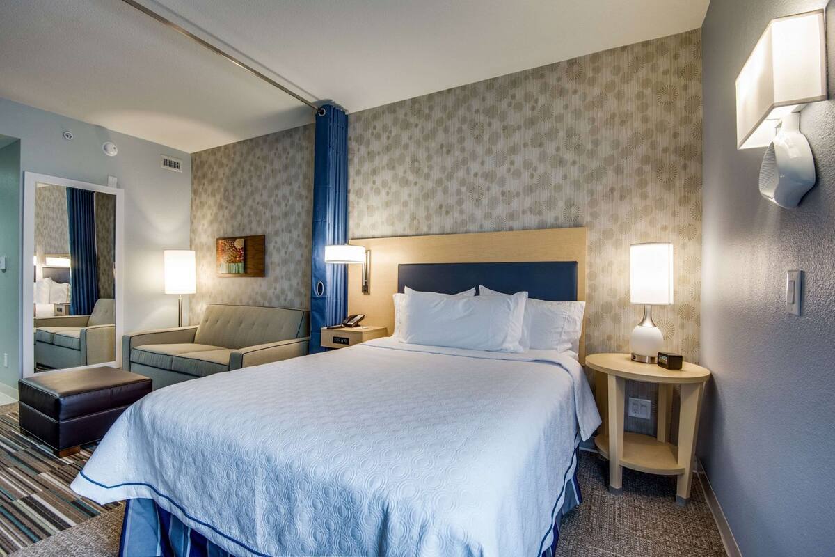 Una habitación de hotel con una cama grande, dos mesitas de noche con lámparas, un sofá, una pequeña mesa y un espejo.