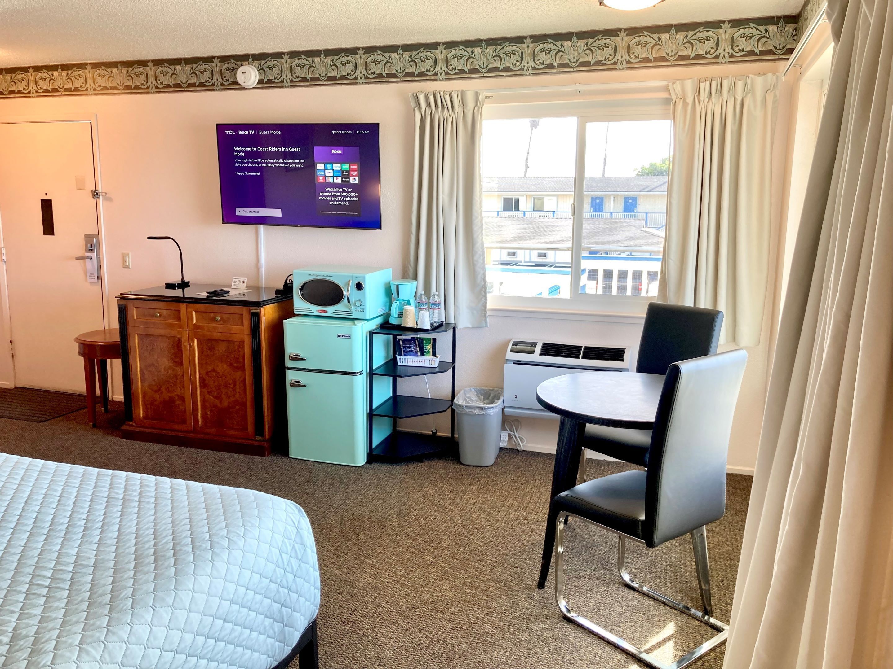 A hotel room with a bed, a desk with a computer, a coffee maker, a TV, and a window with curtains.