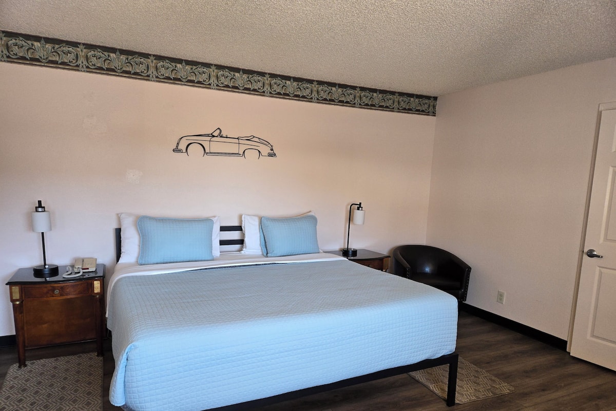 Una cama bien hecha con cubrecama acolchado, una mesita de noche con lámpara, una silla y un adorno de pared con motivo de coche.