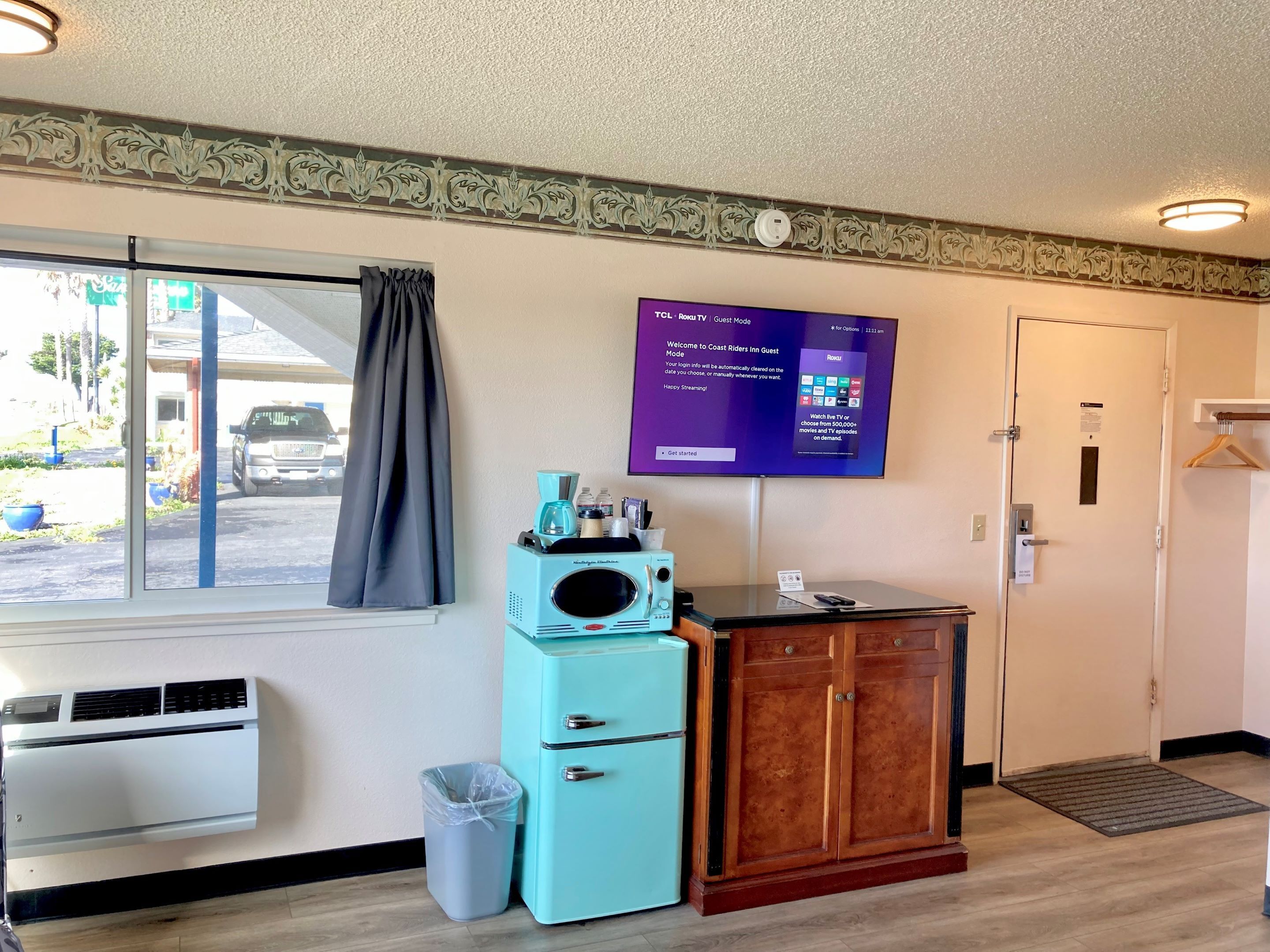 A hotel room with a large window, a flat-screen TV mounted on the wall, a small kitchenette with a coffee maker, a trash can, and a wooden cabinet.