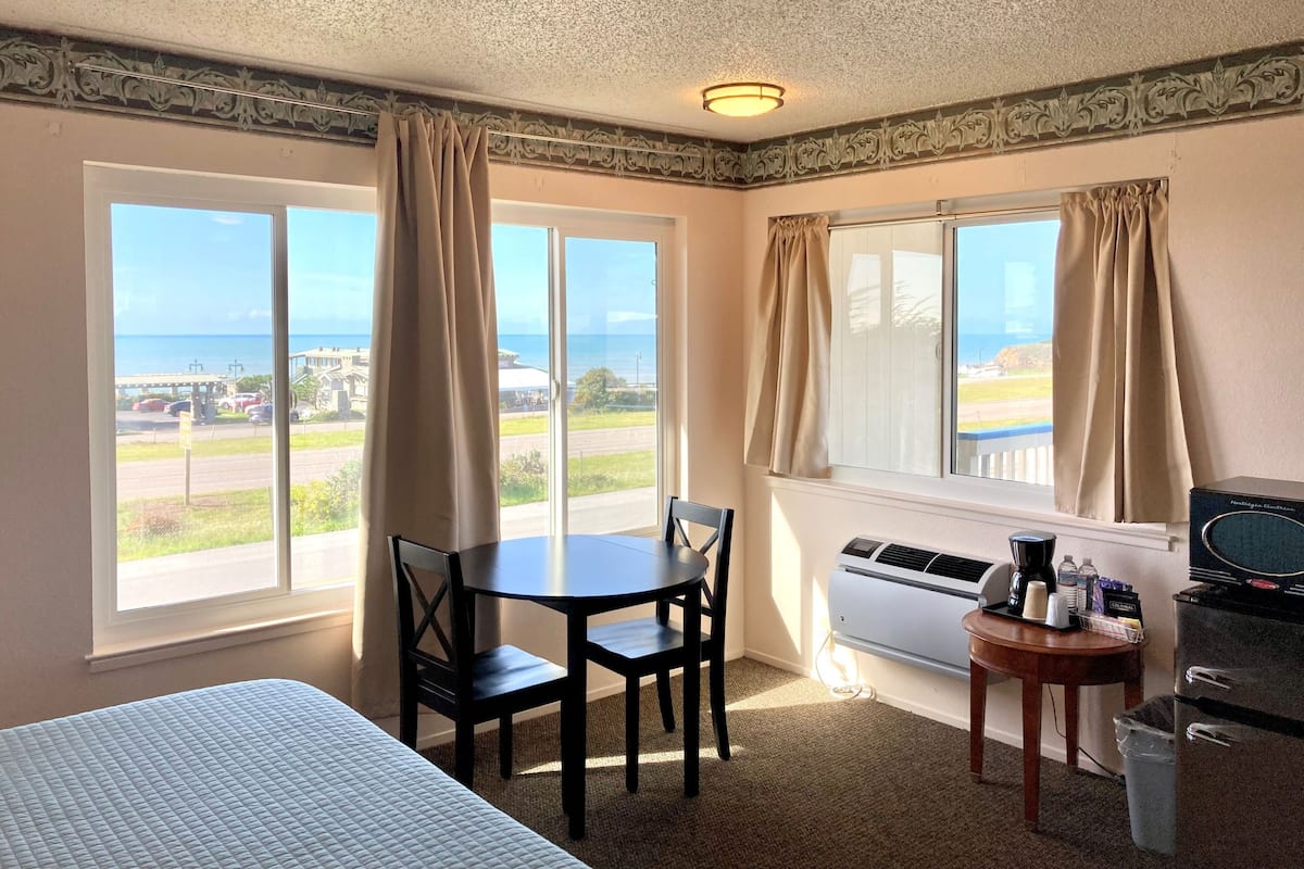 Habitación de hotel con cama, una mesa con sillas, un escritorio con computadora, unidad de aire acondicionado y ventanas amplias con cortinas.