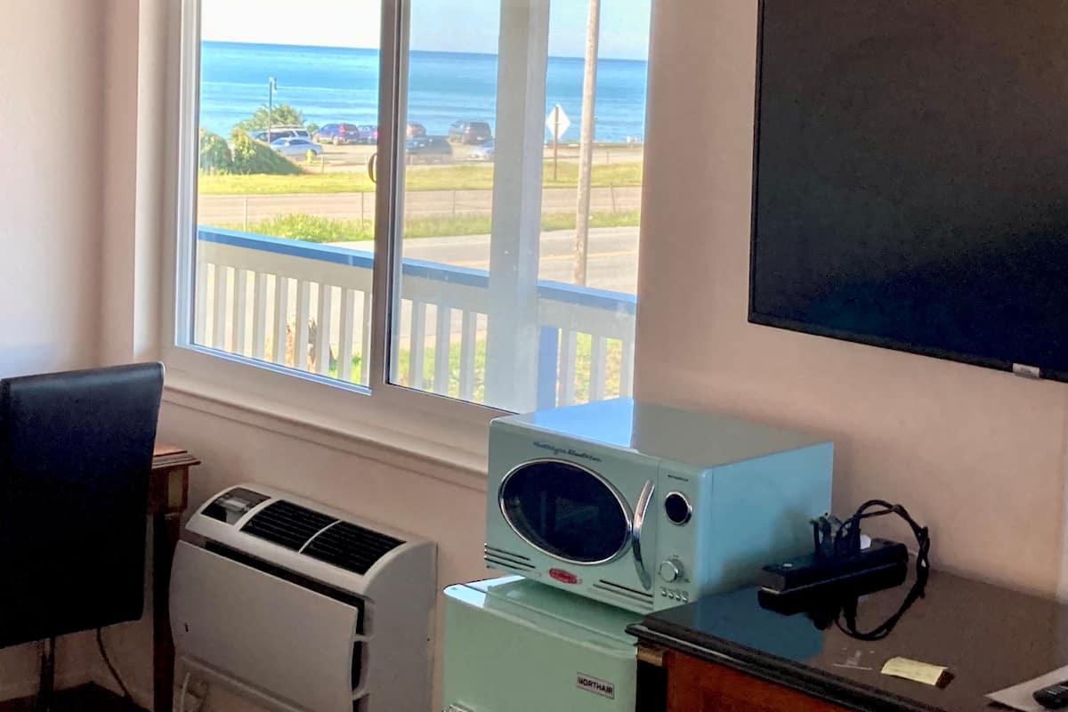 Una habitación con un amplio ventanal que da a una playa y al océano, un aire acondicionado blanco, un microondas de estilo vintage y un televisor de pantalla plana montado en la pared.
