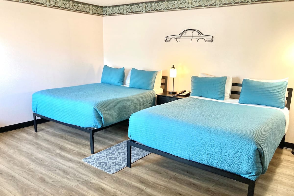 Habitación de hotel con dos camas, cada una con una colcha azul y almohadas a juego. Entre las camas, una mesita de noche con una lámpara y un teléfono.