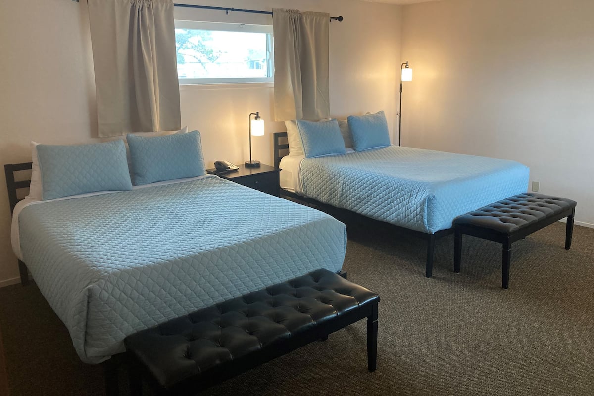 Habitación de hotel con dos camas, cada una con cubrecama azul acolchado y almohadas a juego, un banco, lámparas de noche y una ventana con cortinas.