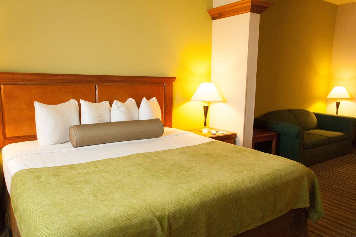 Une chambre d’hôtel avec un grand lit, deux lampes de chevet, un fauteuil vert et une tête de lit en bois.